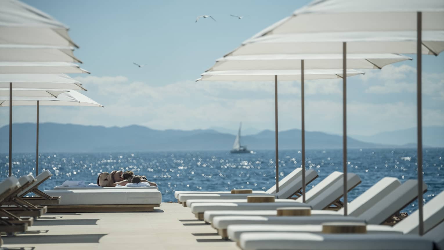 A sail boat sails in the distance while white lounge chairs are ready for guests and lined up next to the oceanside Nafsika Pool in Athens. 