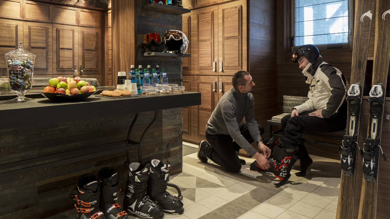 Ski concierge fits boots on man sitting on bench by skis, table with fresh fruit water