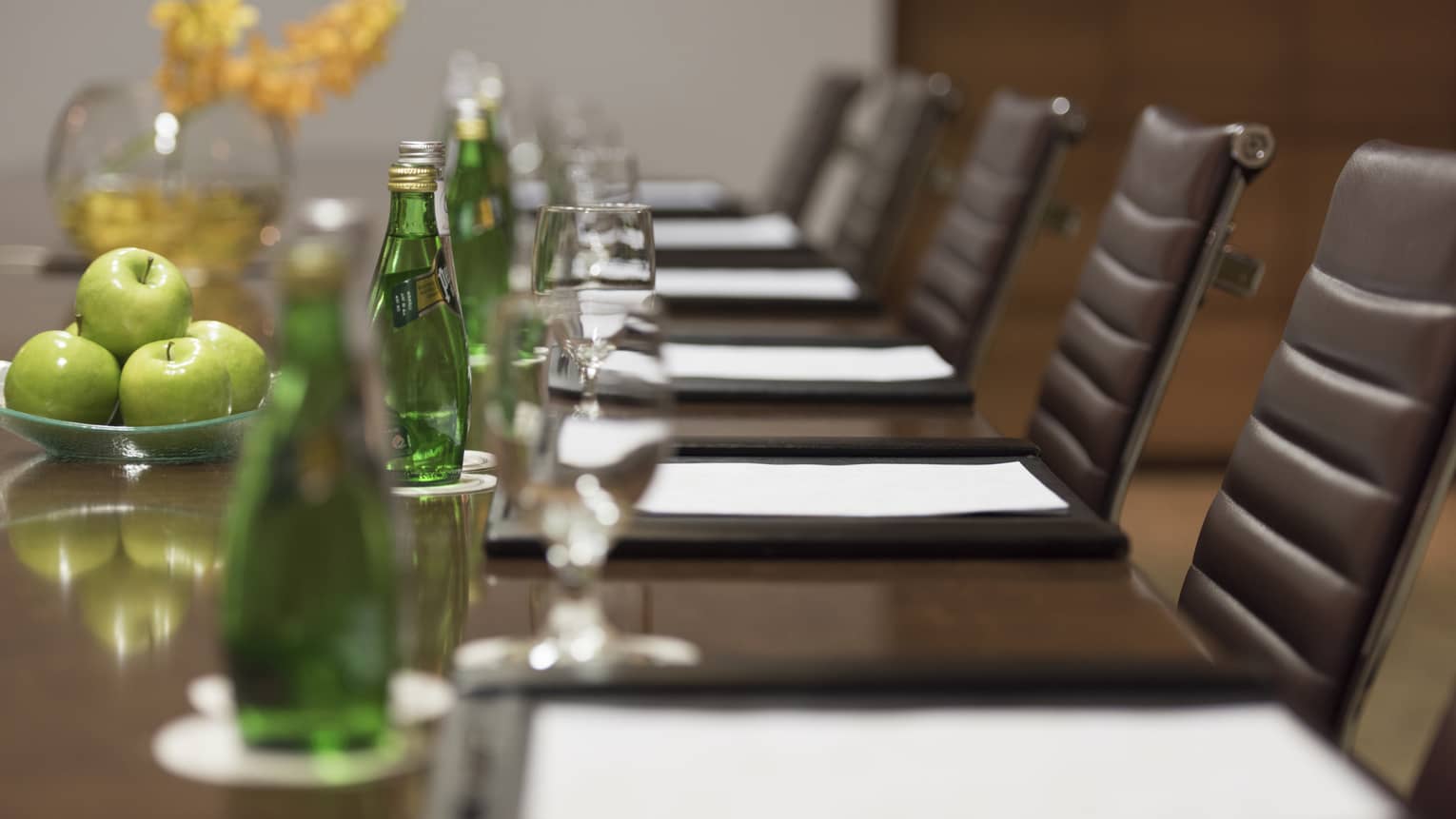 Close-up of business meeting table row of chairs, paper agendas, green glass bottles, apples