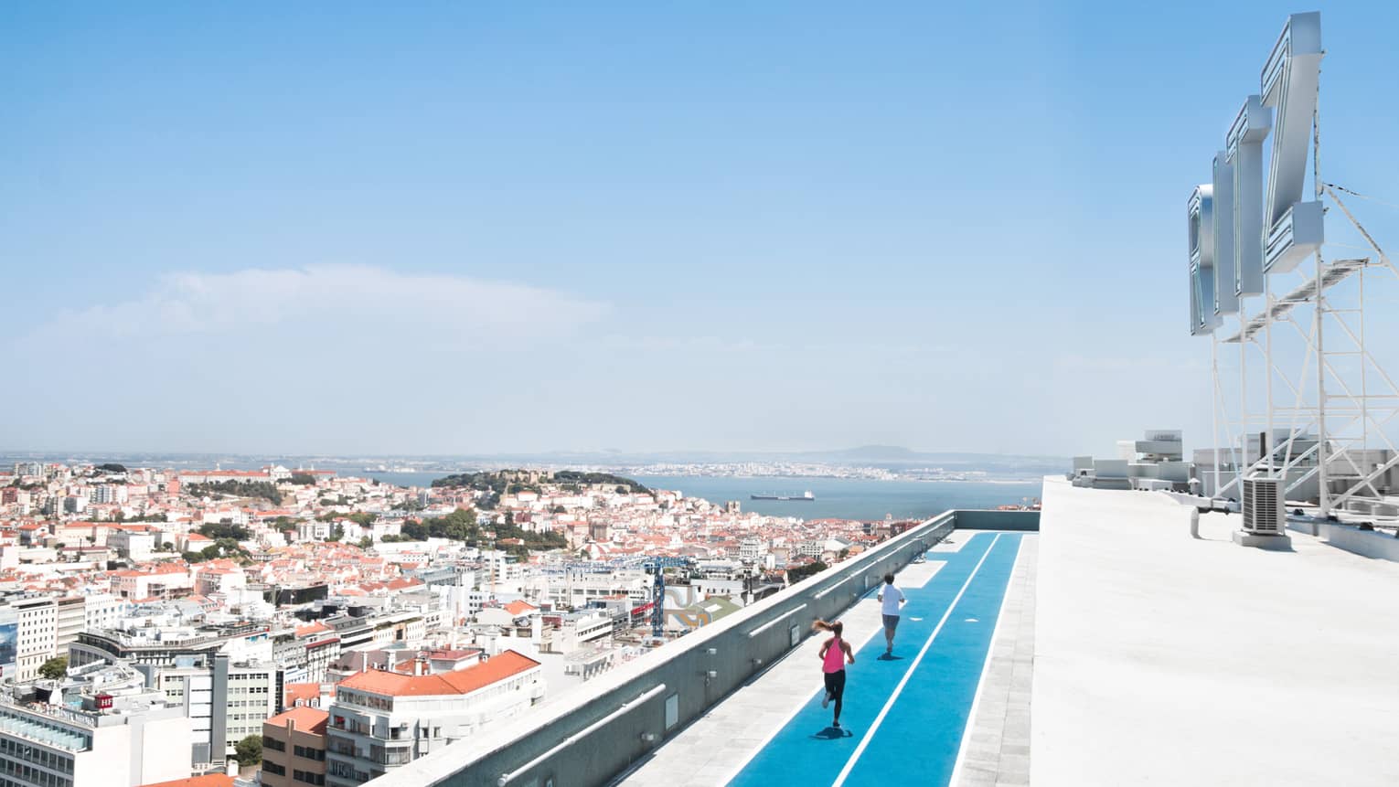 Aerial view of backs of man and woman running on blue rooftop outdoor track, Lisbon city roofs below