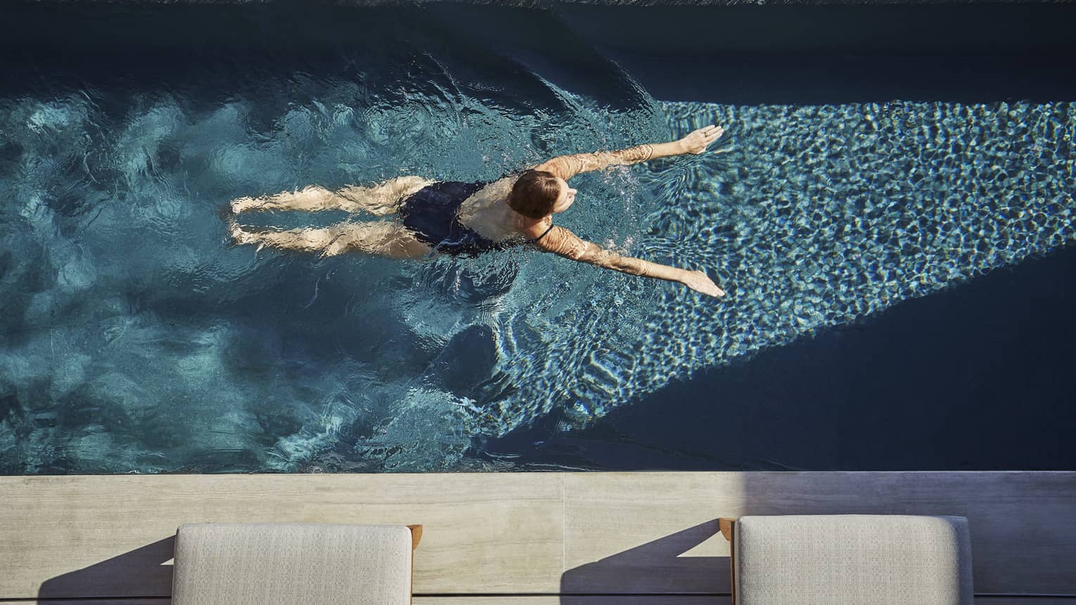 Woman in dark bathing suit swimming in pool. Pool deck and lounge chairs visible.