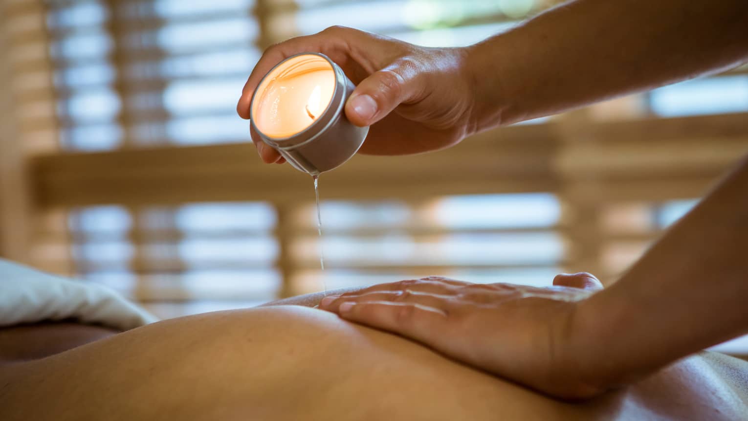 Massage therapist pours hot wax from tealight candle over person's bare back in spa