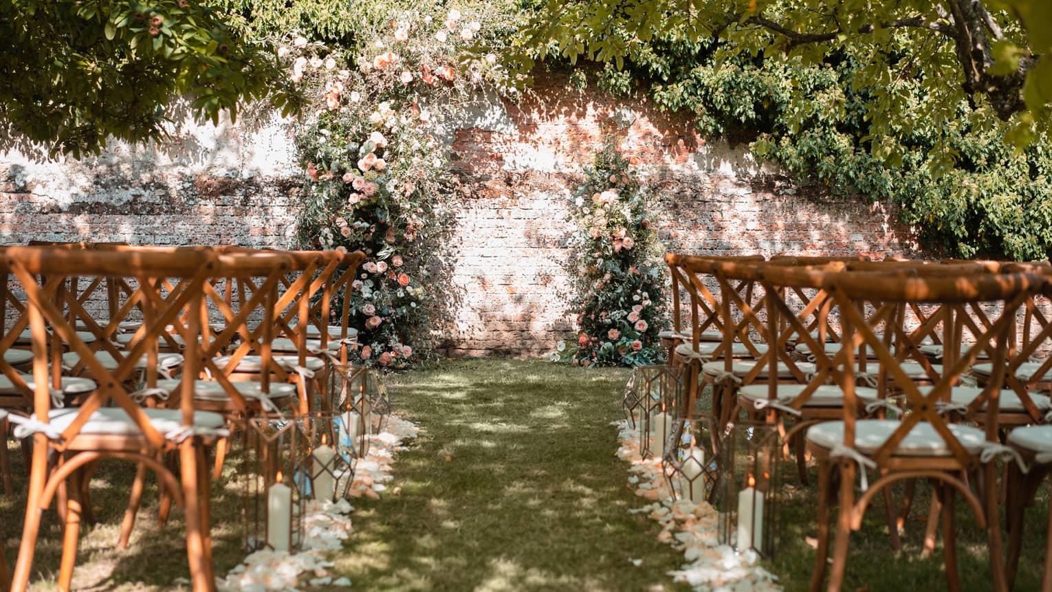 Outdoor wedding aisle lined with flower petals and chairs leading to a brick wall draped with greenery