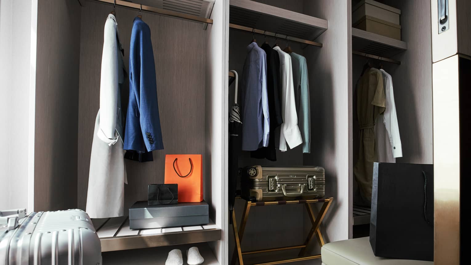 Hotel closet with hanging clothes, including blazers and shirts, along with suitcases, shopping bags and storage boxes on shelves
