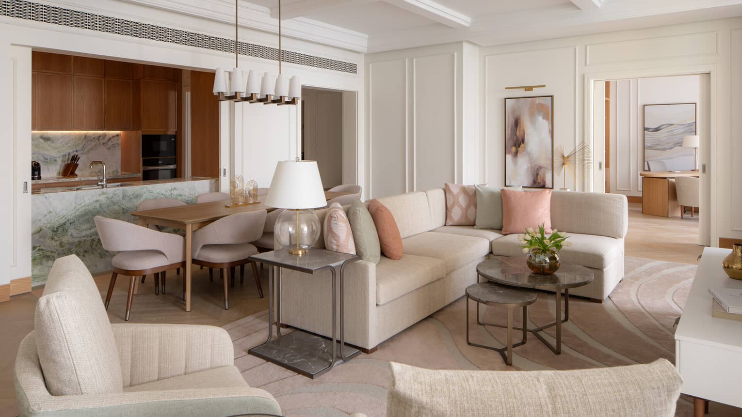 Elegant open-plan living space with neutral furnishings, pastel accents, a marble kitchen island and a dining area beside a modern chandelier.