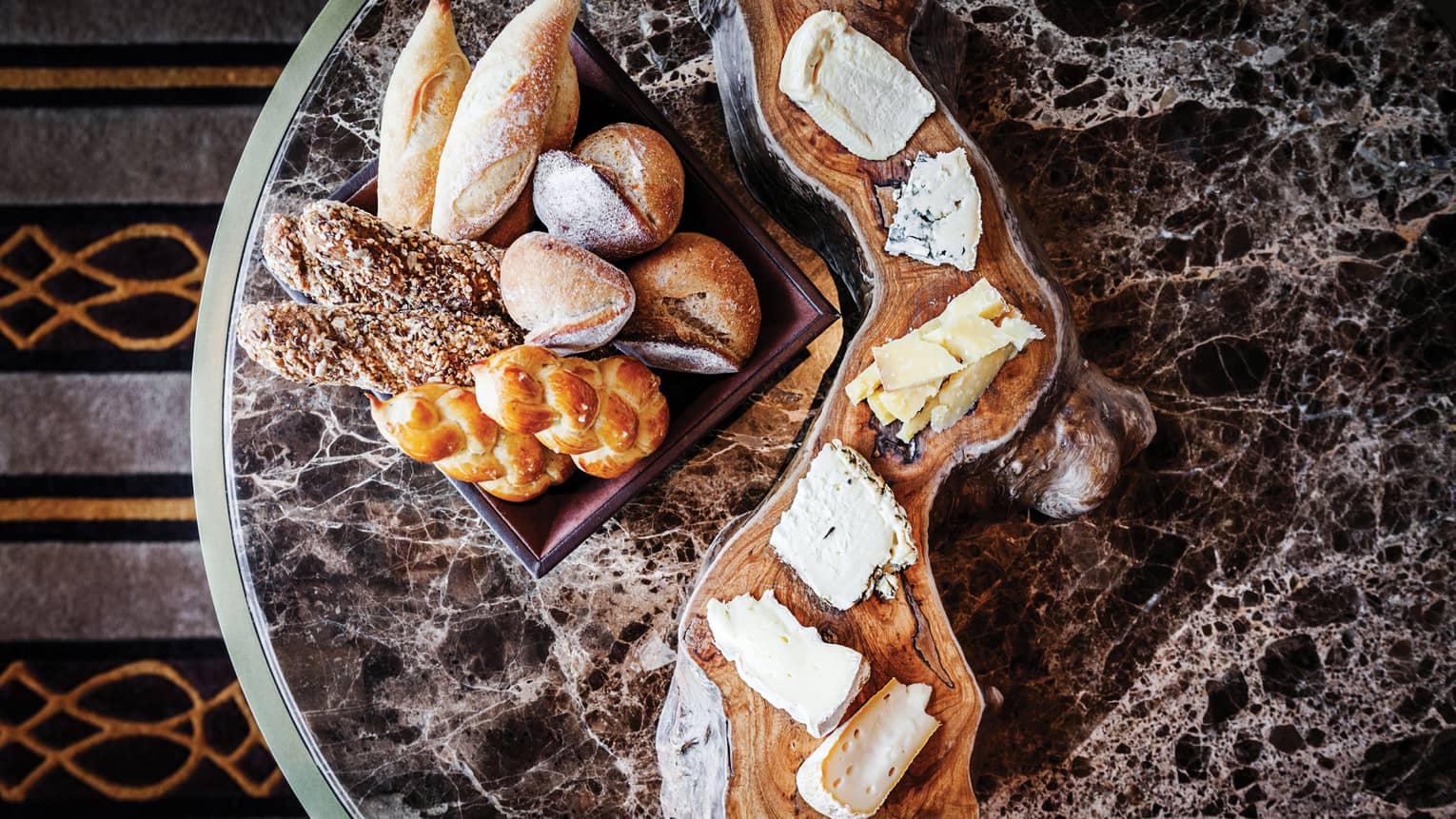 Assorted breads in a basket and a selection of cheeses on a rustic wooden board, set on a marble table.