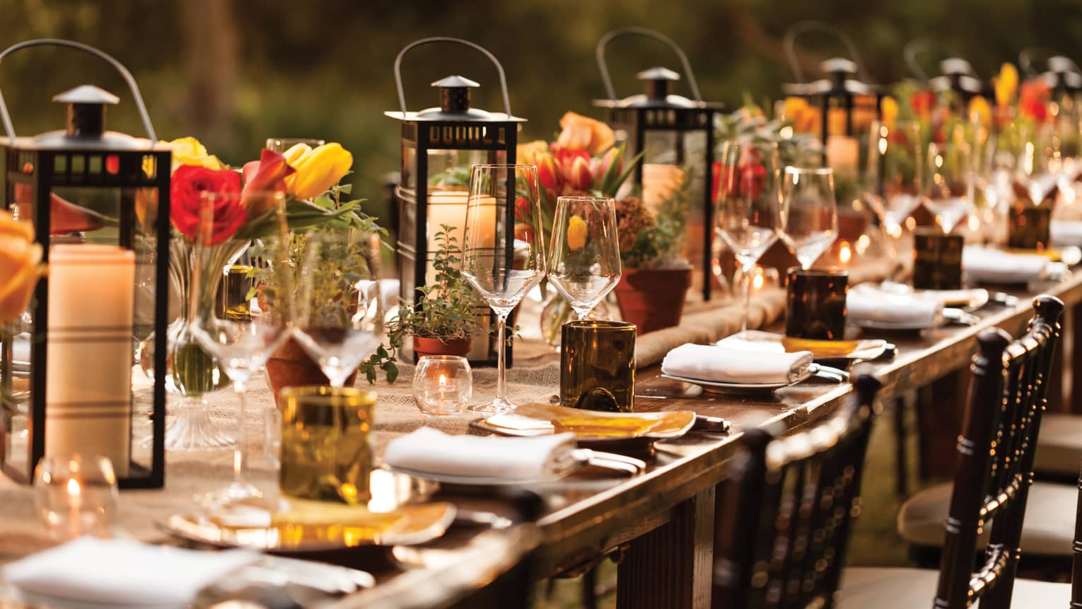 Close-up of long outdoor dining table set with lanterns, candles, wine glasses, flowers