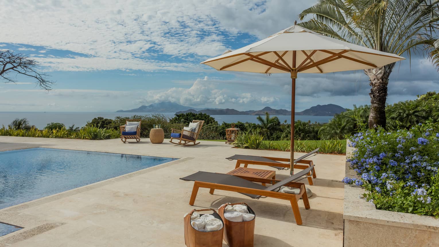 Two lounge chairs under patio umbrella, baskets with towels by outdoor pool