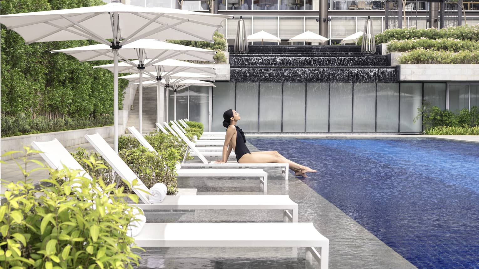A woman in a black bathing suit relaxes on a chaise by the pool