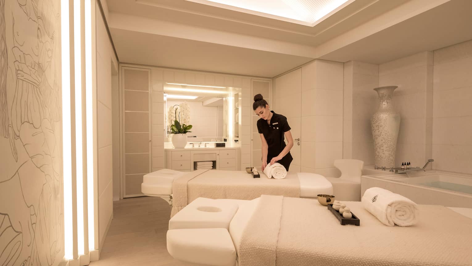 Spa staff rolls white towel on couples massage beds in white treatment room
