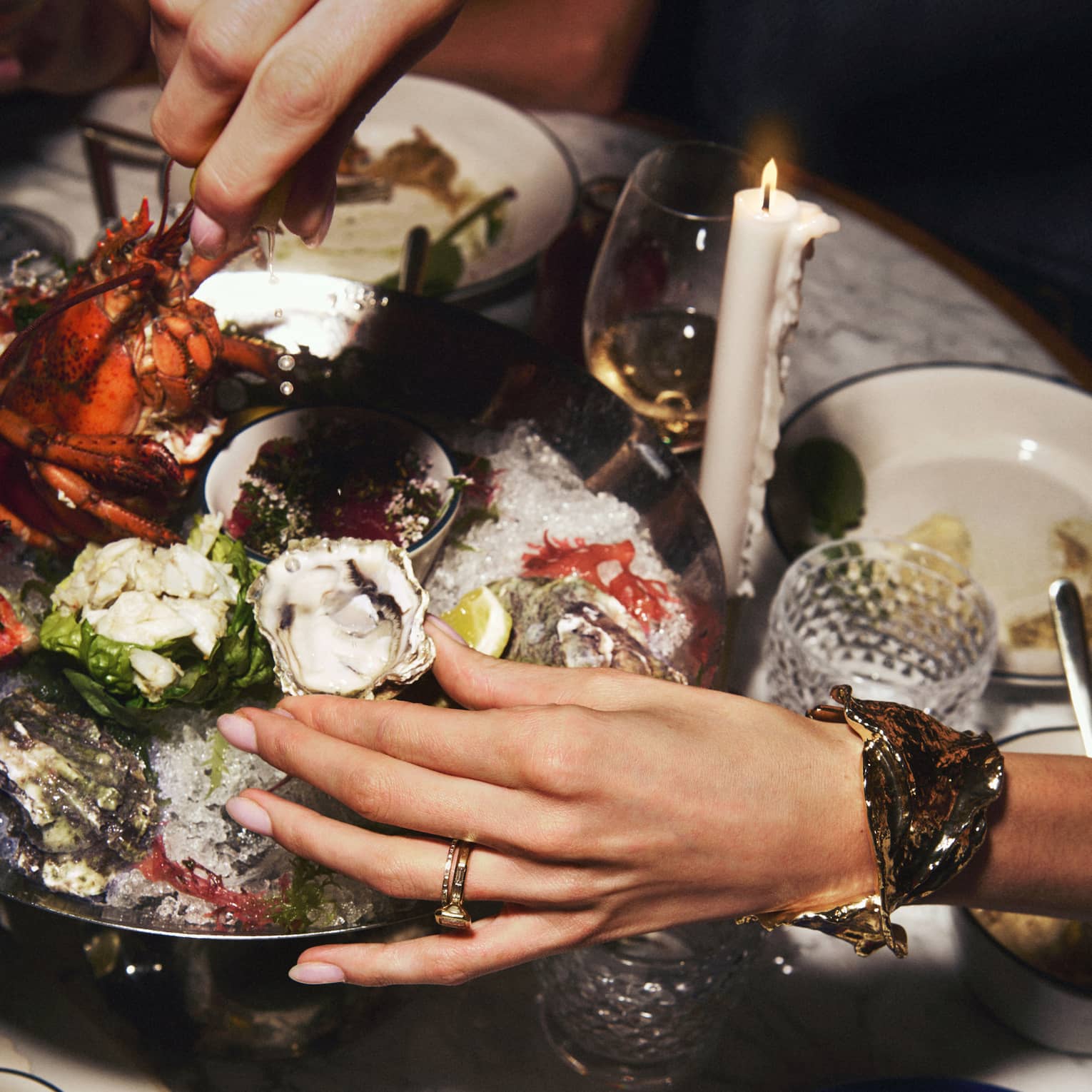 A pair of hand reaches to grab an oyster from a silver seafood platter