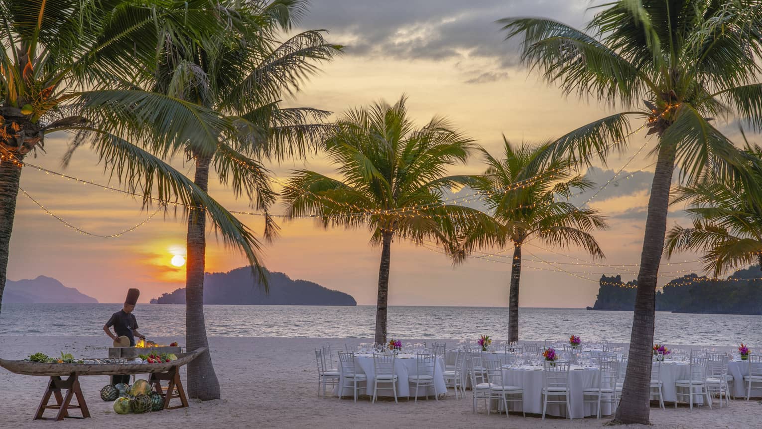 Tables set for dinner on the beach under a canopy of palm trees, a chef cooking over an open flame against the setting sun.