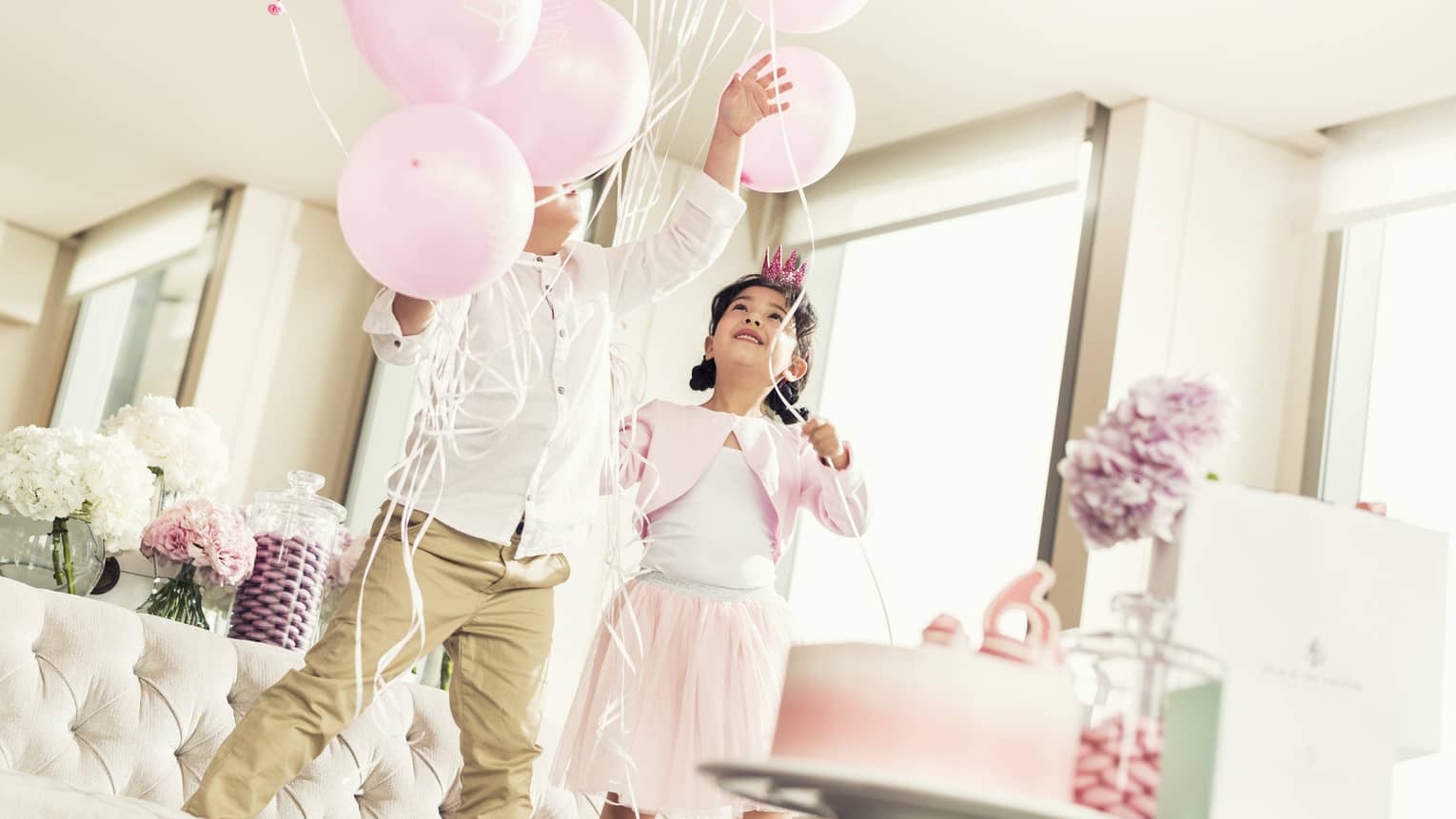Two kids playing with pink balloons near a pink cake.