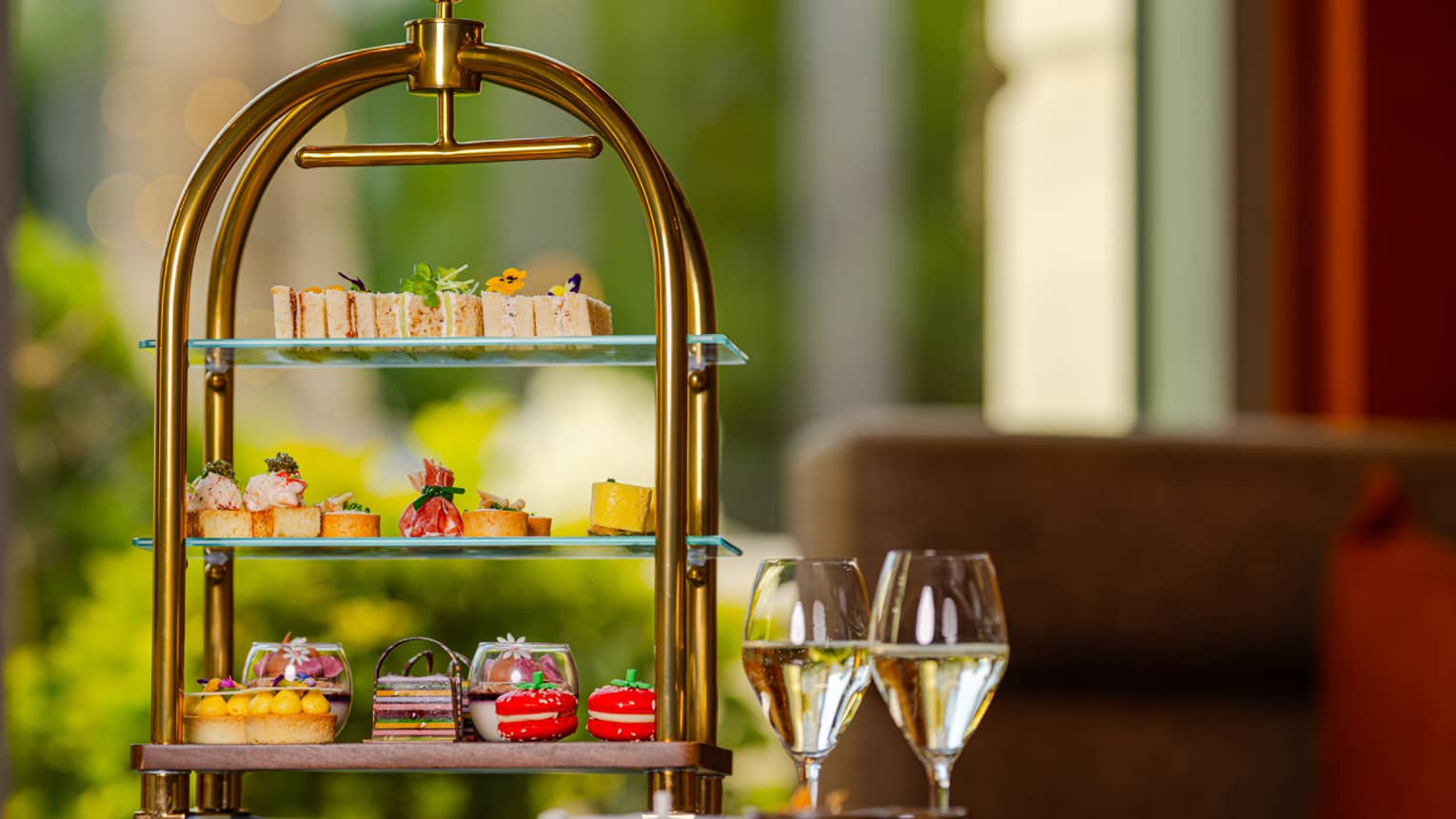 A tiered serving tray holds a variety of colourful small bites and is sitting next to two stemmed glasses containing light-hued liquid.