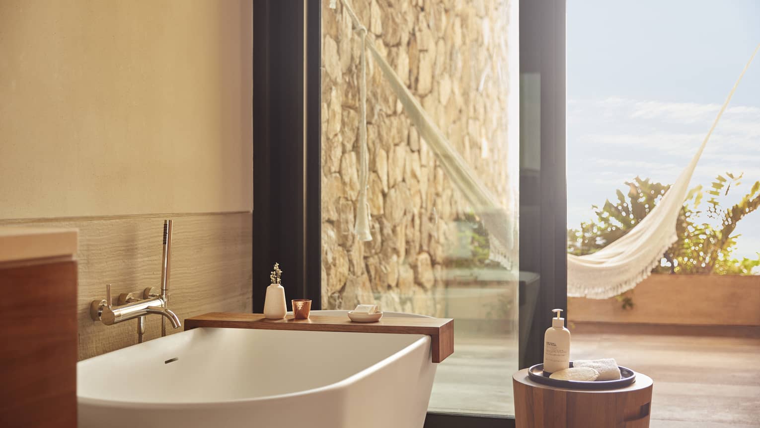 Serene bathroom with a modern white bathtub, wooden accents and glass doors opening to an outdoor area with a hammock and stone wall, bathed in natural light