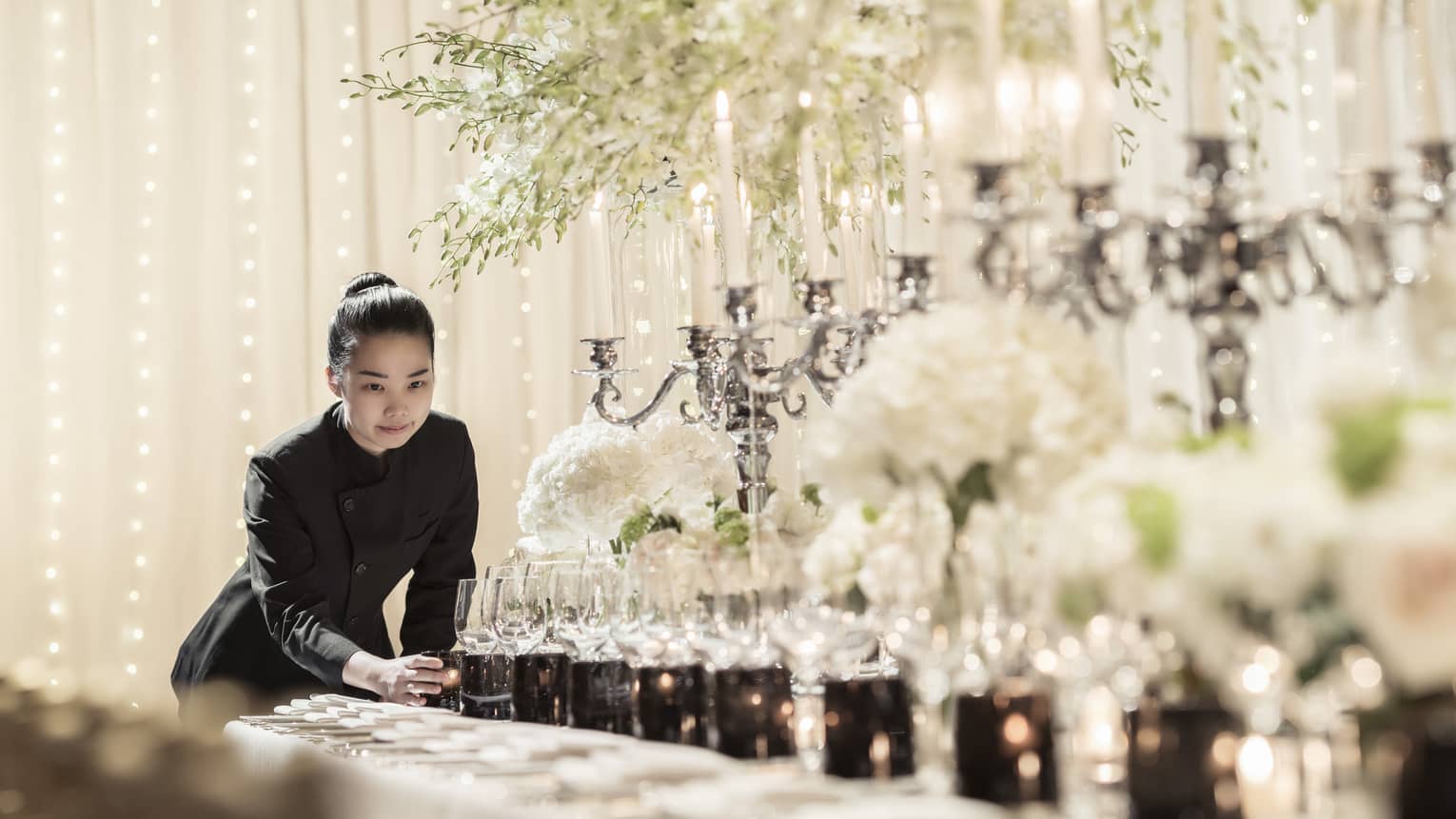 Hotel staff places candle at end of long formal dining table set with candelabras, sparkling lights