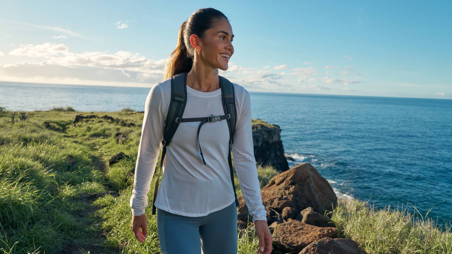 Close-up of a smiling, backpacked guest standing atop a grassy coastal cliff, clear sky and ocean with gentle wake beyond.