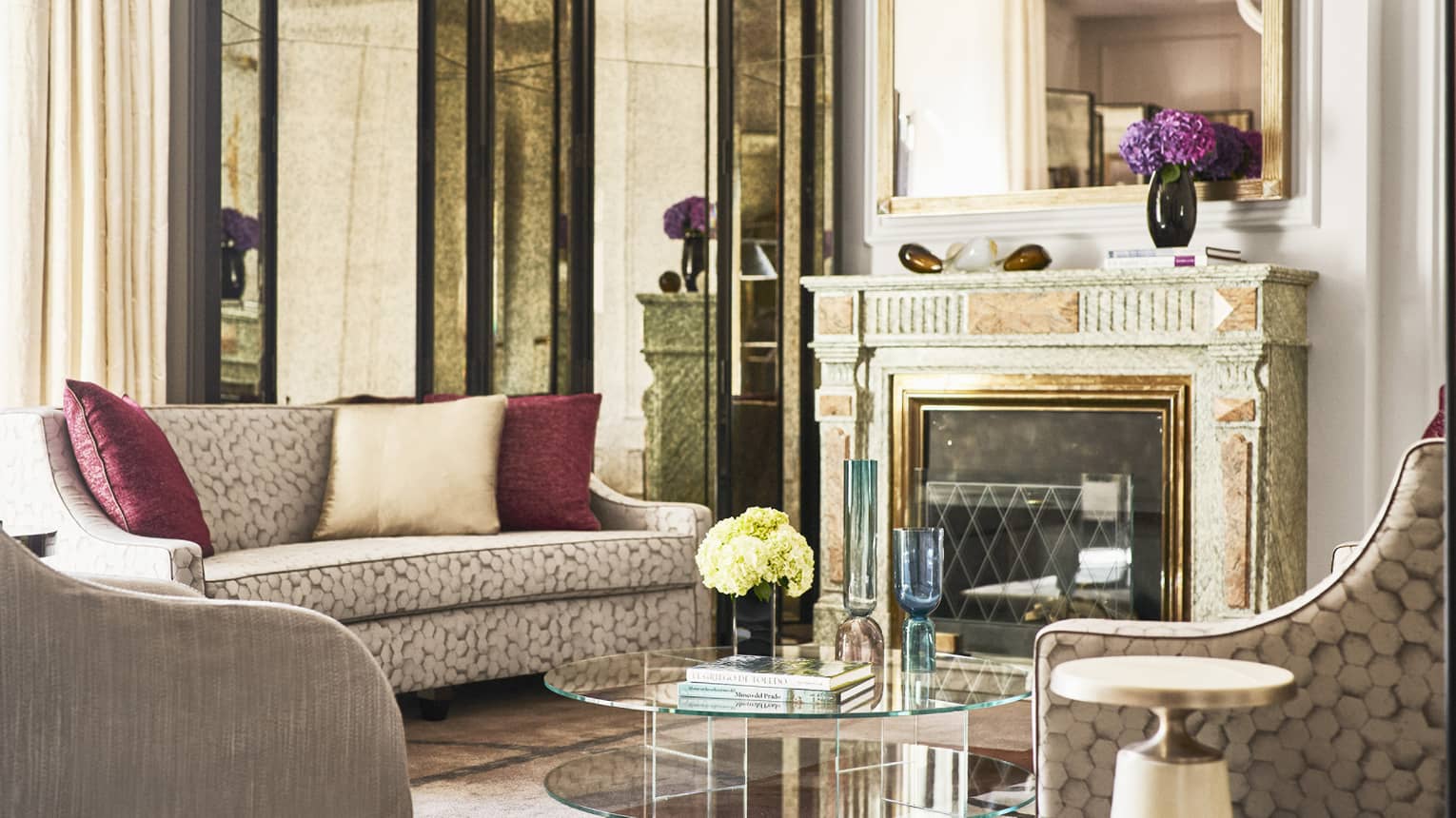 Luxury hotel suite with patterned sofa with red cushions, armchairs, glass coffee table with flowers and books and ornate fireplace with large mirror above