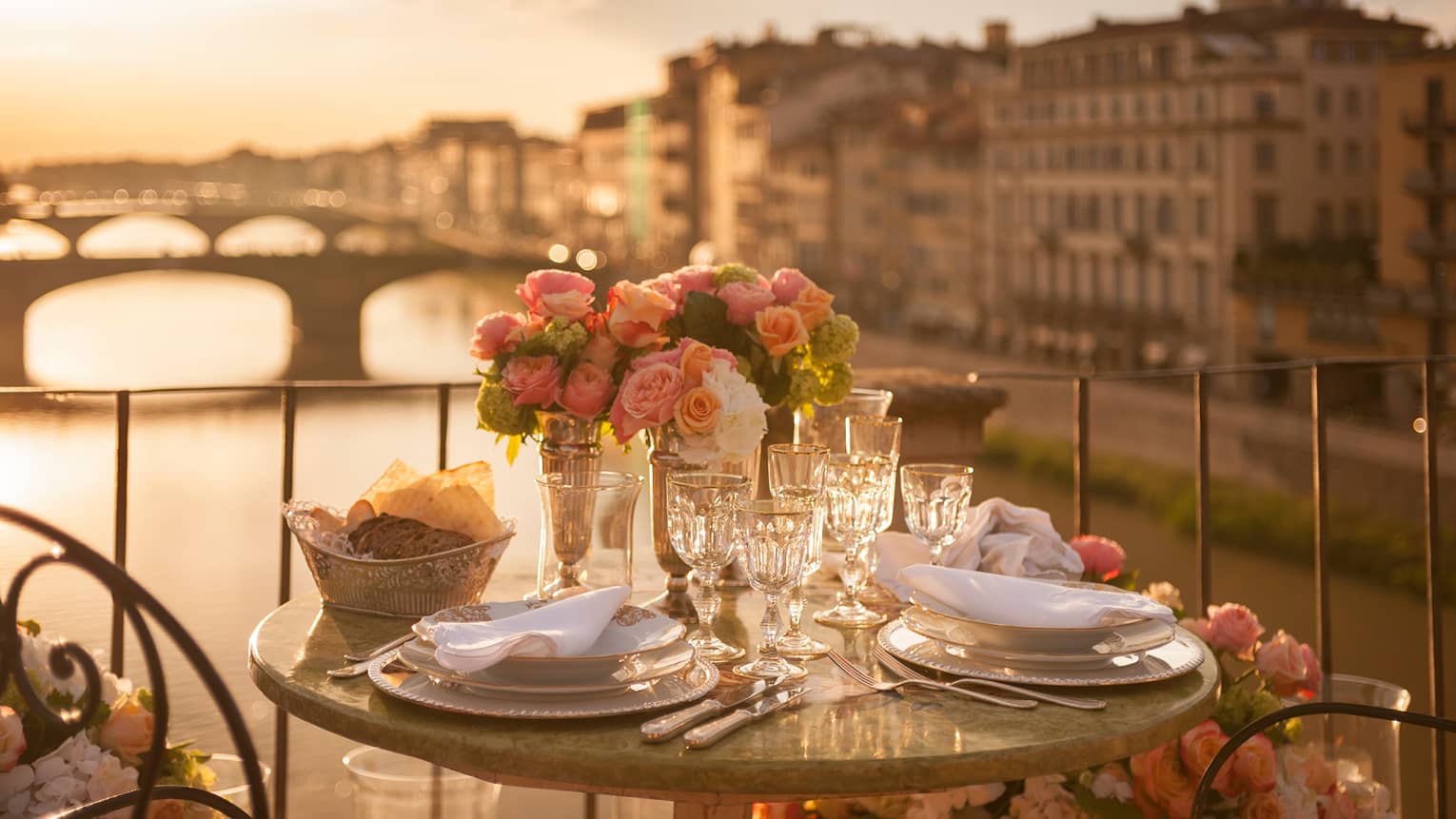 A romantic table for two is set with white porcelain plates, crystal glasses, pink roses on a balcony overlooking a bridge and historic building in Florence