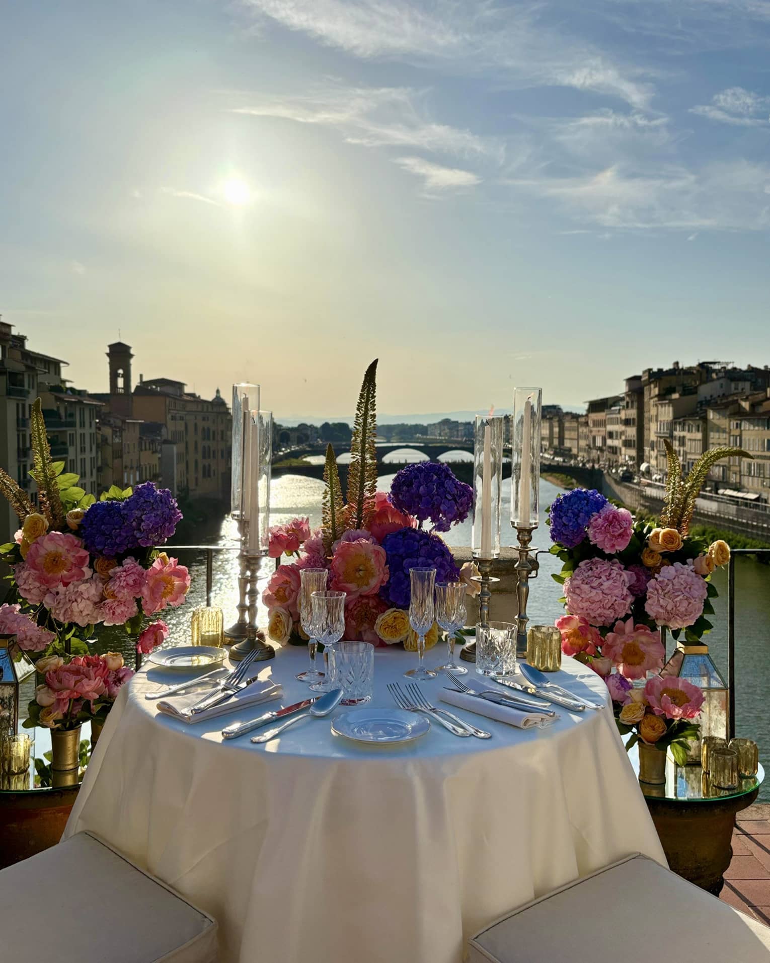 An elegantly set outdoor dining table overlooking a a river, with a sunny sky in the background.