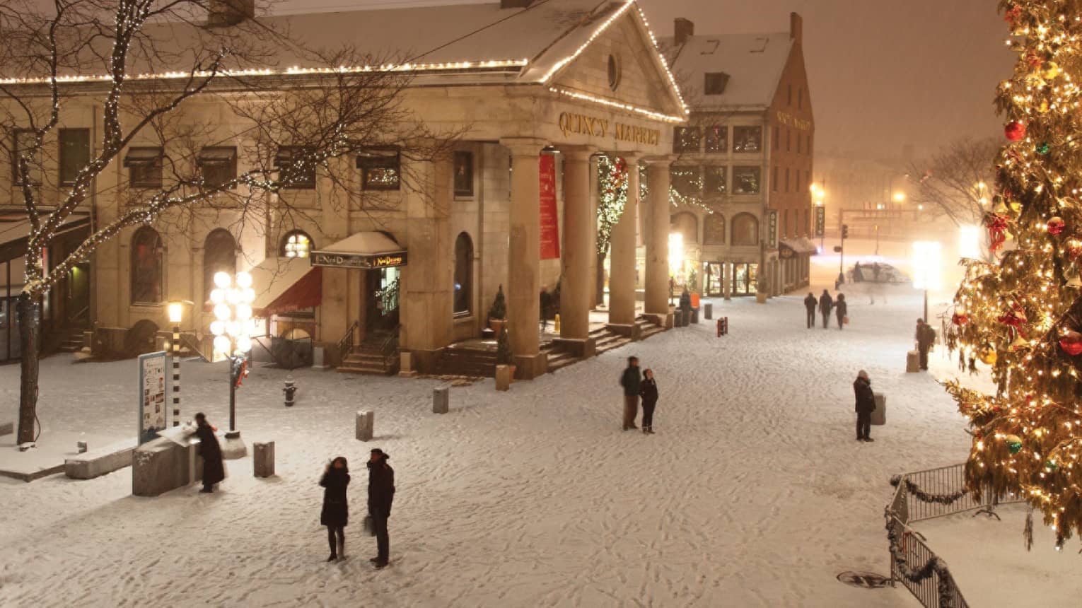 A snowy scene in boston, near historic buildings across from a large well-lit christmas tree