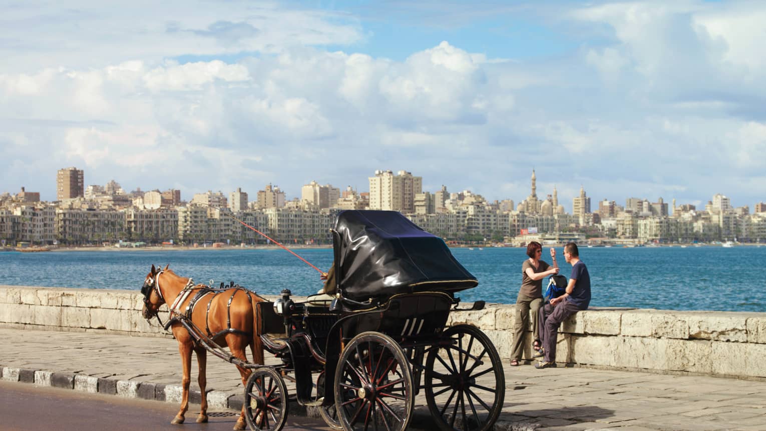 Caleche on the Corniche with couple seated on low rock wall, overlooking water and city skyline