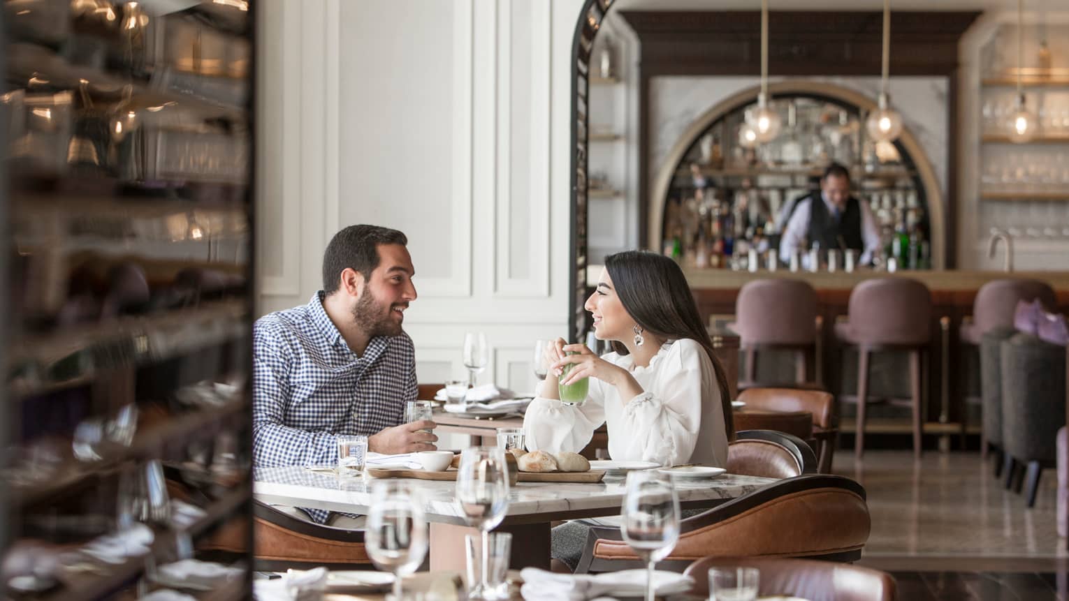 Smiling man and woman sit at sunny dining table with colourful drinks and bread, bartender in background