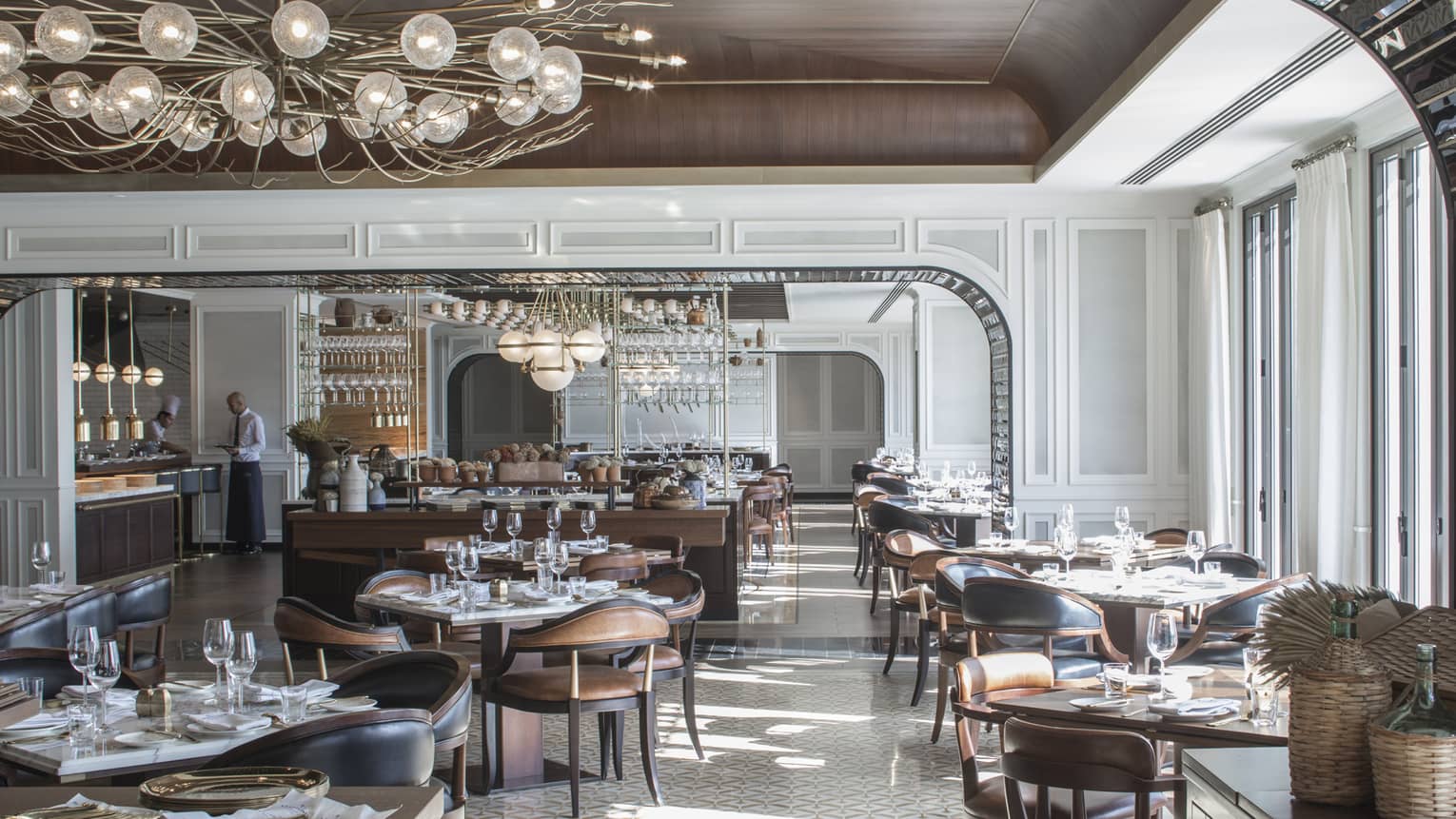 Large, sunny La Capitale dining room with elegant brown leather chairs, tables, white tile, modern chandeliers, server at counter