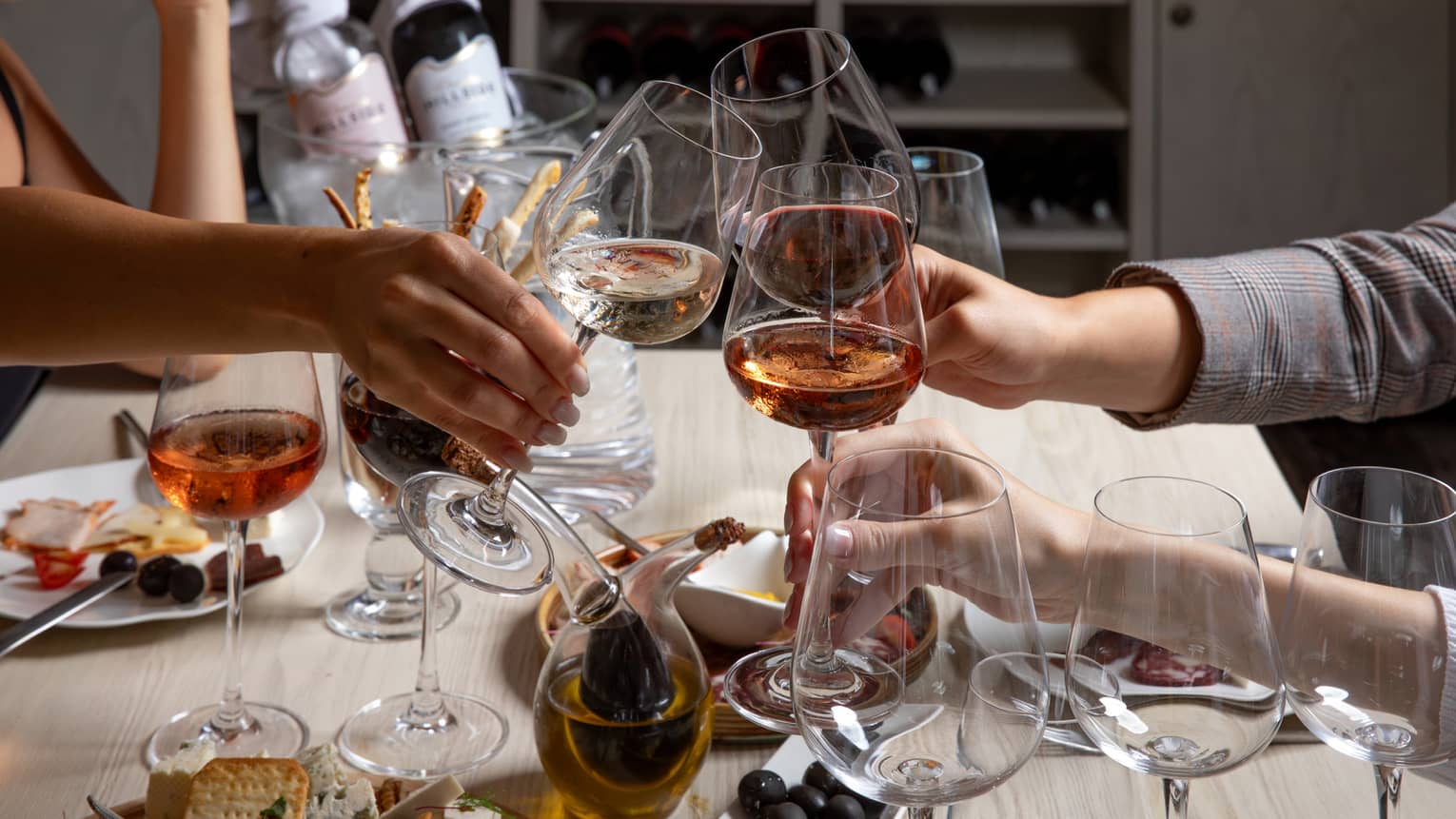 Three wine enthusiasts clink glasses over a table of goblets, appetizers and an ice bucket containing two bottles of wine.