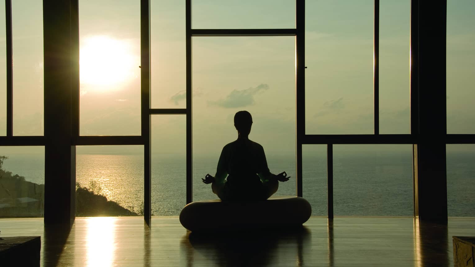 Silhouette of person meditating on cushion in front of floor-to-ceiling windows at sunset