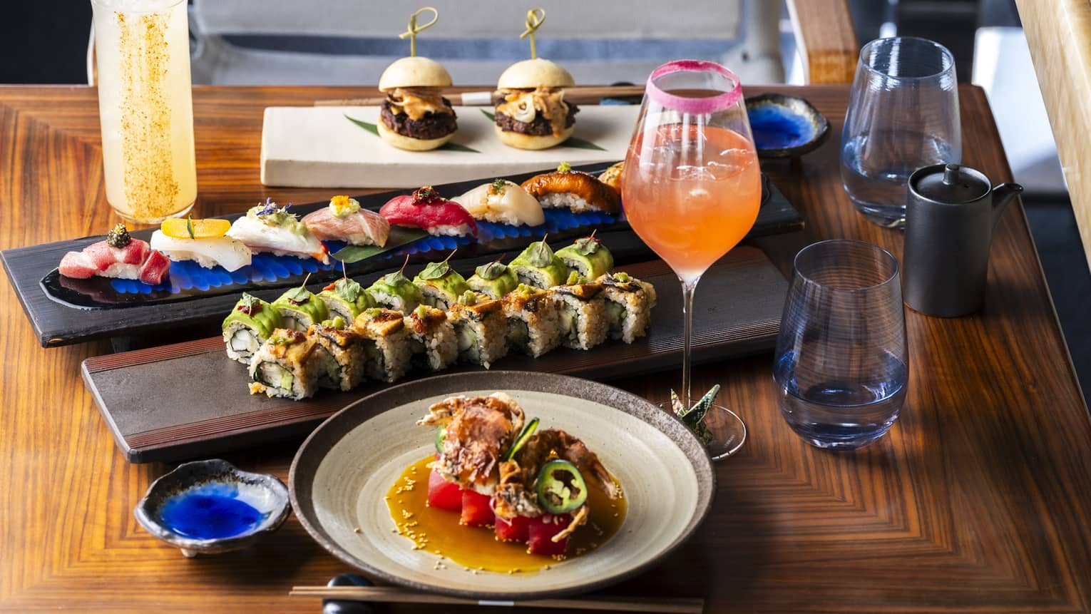 A wood table holds rectangular and round plates with sushi and other small bites. Colourful drinks in stemware and tumblers are next to the plates.