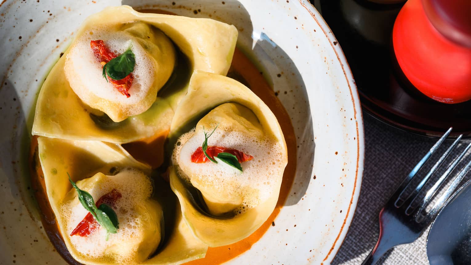 Aerial view of three ravioli nested in speckled bowl with green and red garnishes