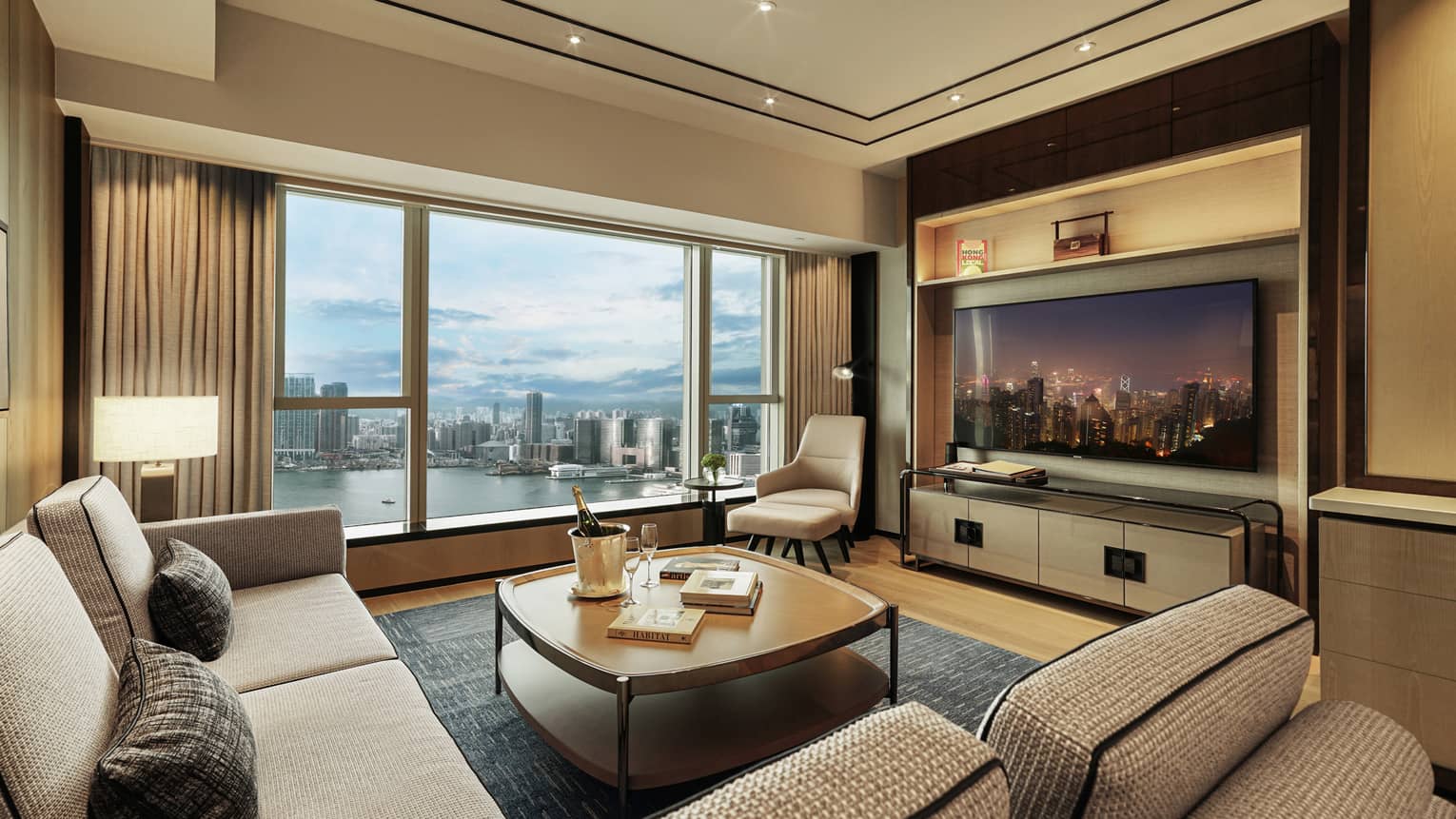 Modern hotel living room with city and harbor view, beige sofas, armchair, TV displaying night skyline and coffee table with champagne.