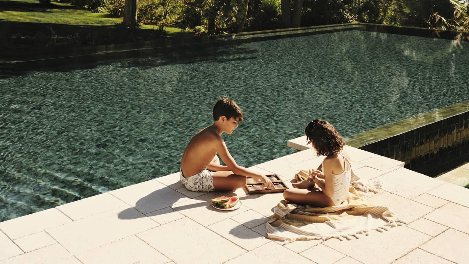 Two seated children play a game on a sunny deck beside a tranquil teal pool flanked by tall trees under a bright blue sky.