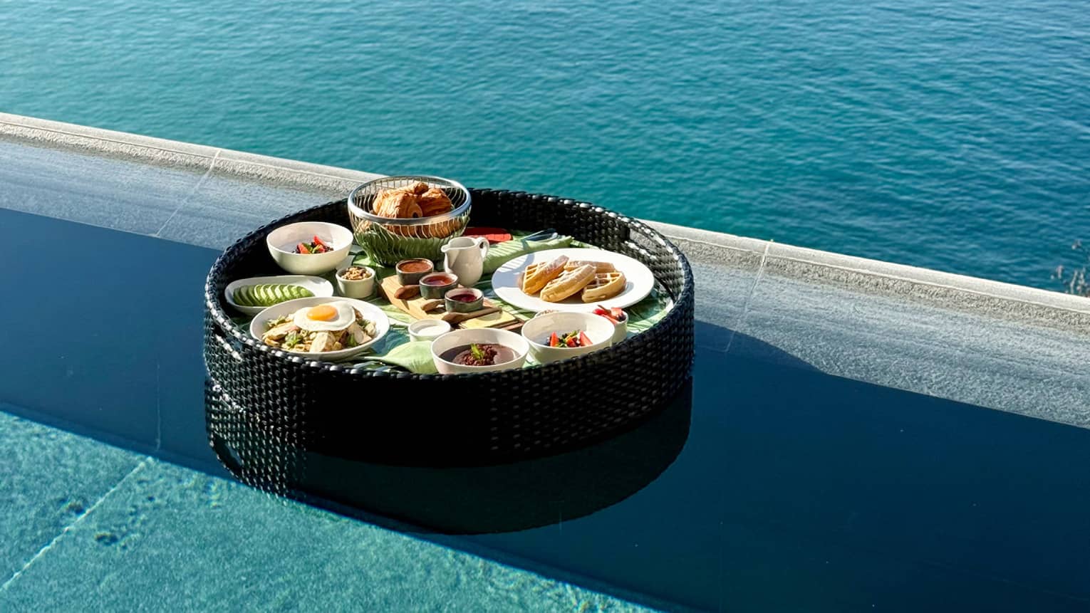A tray of plated breakfast food items on a tray floating near the edge of an infinity pool, with coastline and ocean views.