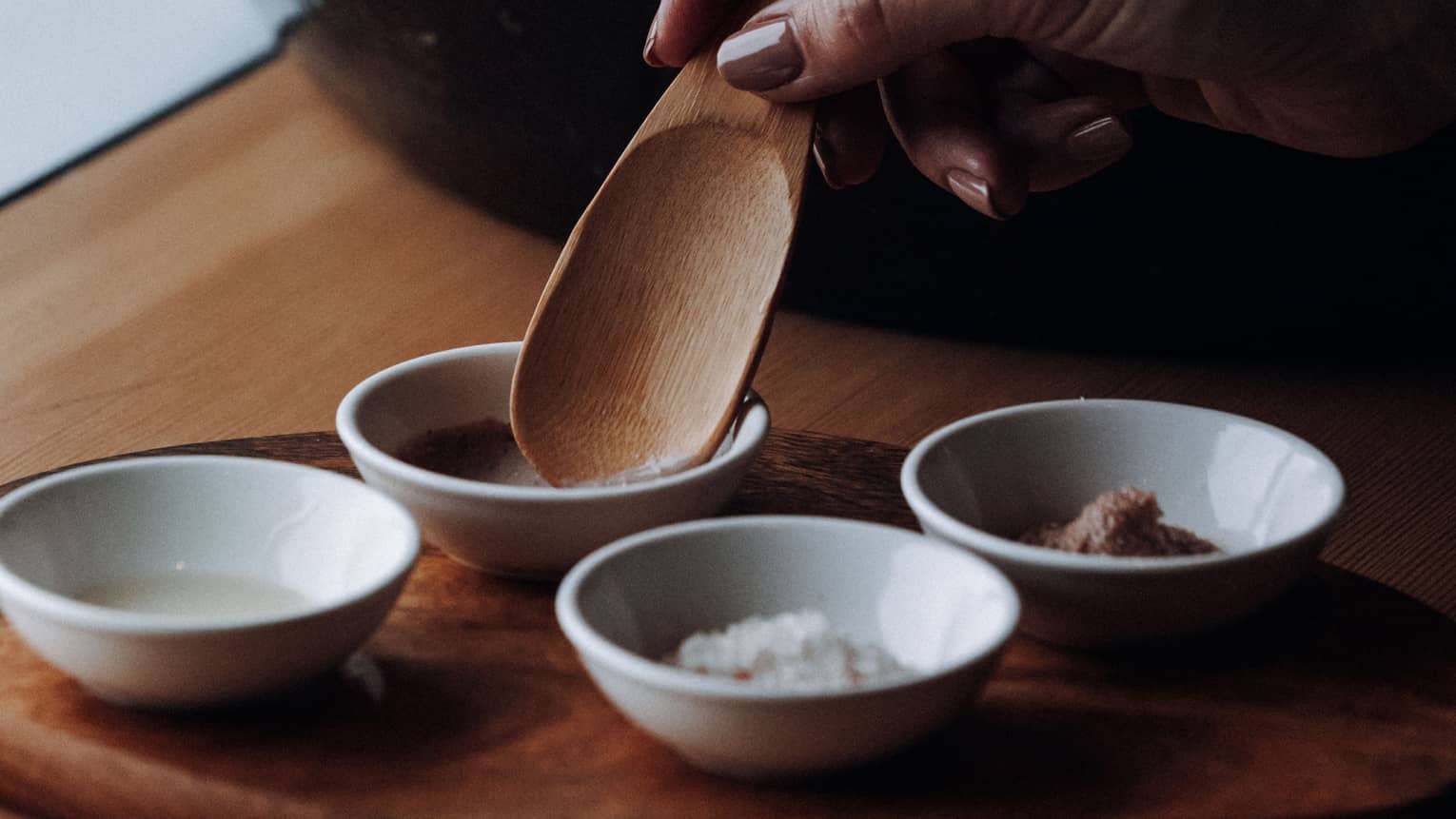 A hand places a wooden spoon into a small clay bowl to scoop up a spa ingredient, with three other clay bowls placed nearby