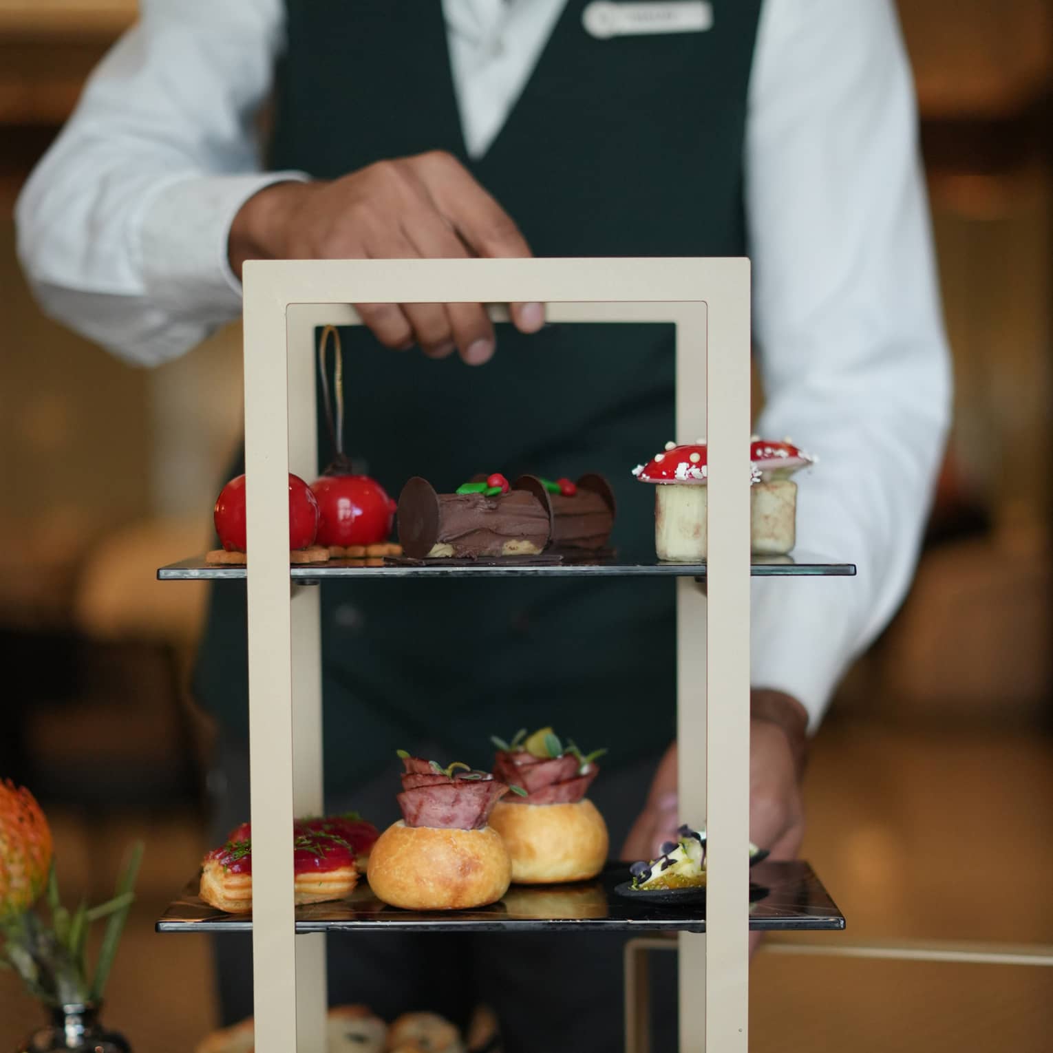 Server wearing black pants, black vest and white button-down shirt presents a three-tiered tray of afternoon tea desserts