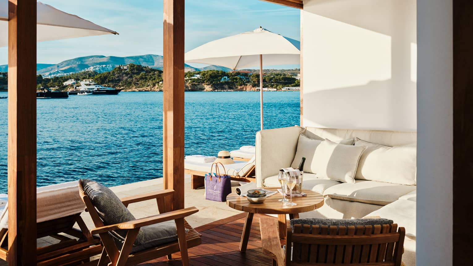 Cabana interior with white banquette-style couch, round teak table and two teak chairs open to platform with white umbrellas and blue sea views