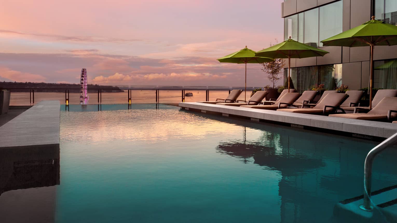 Pink sunset reflected on outdoor swimming pool lined with lounge chairs in front of bay, Seattle Wheel