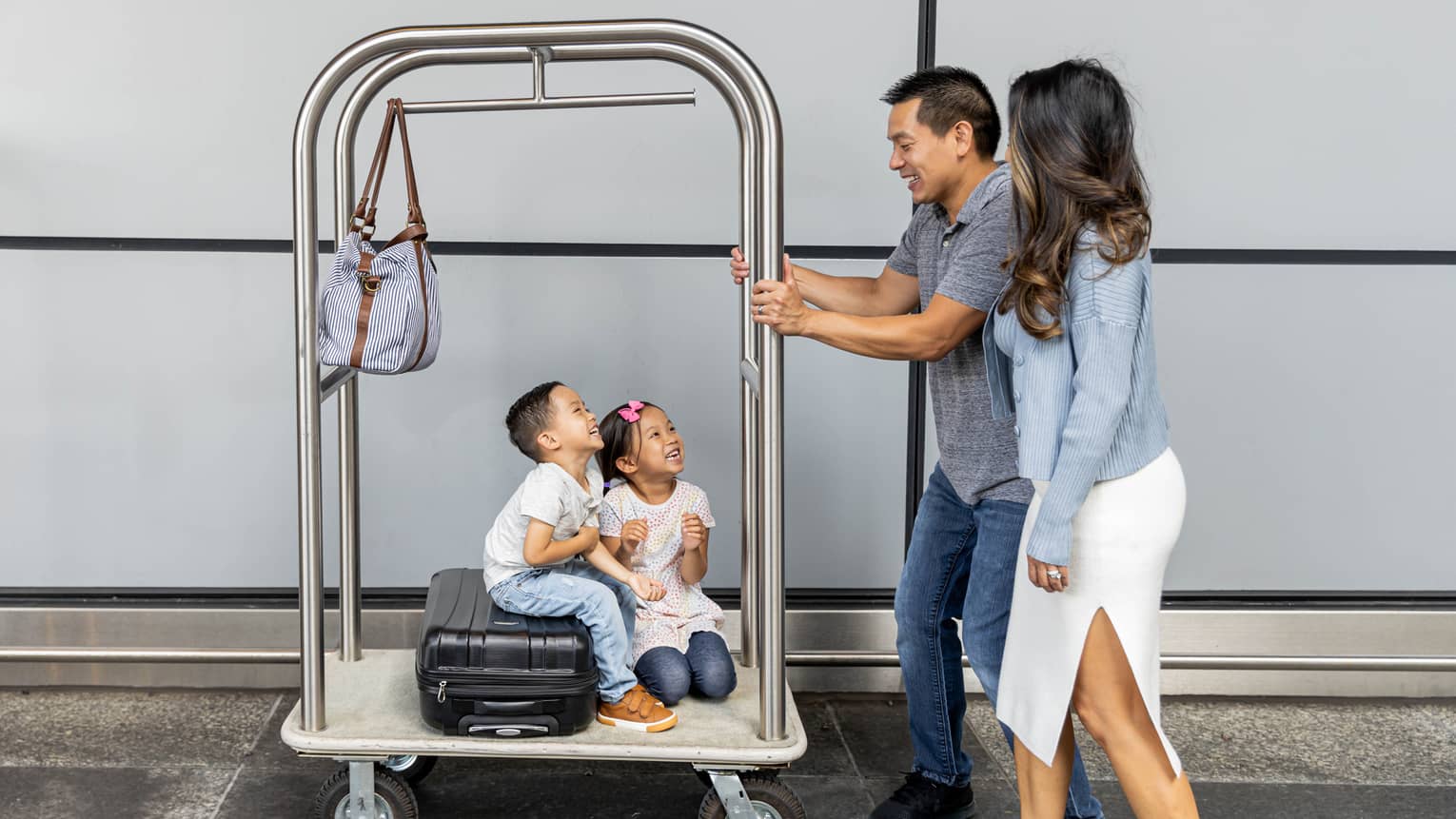 A man and woman pushing a luggage cart with two small children on it.