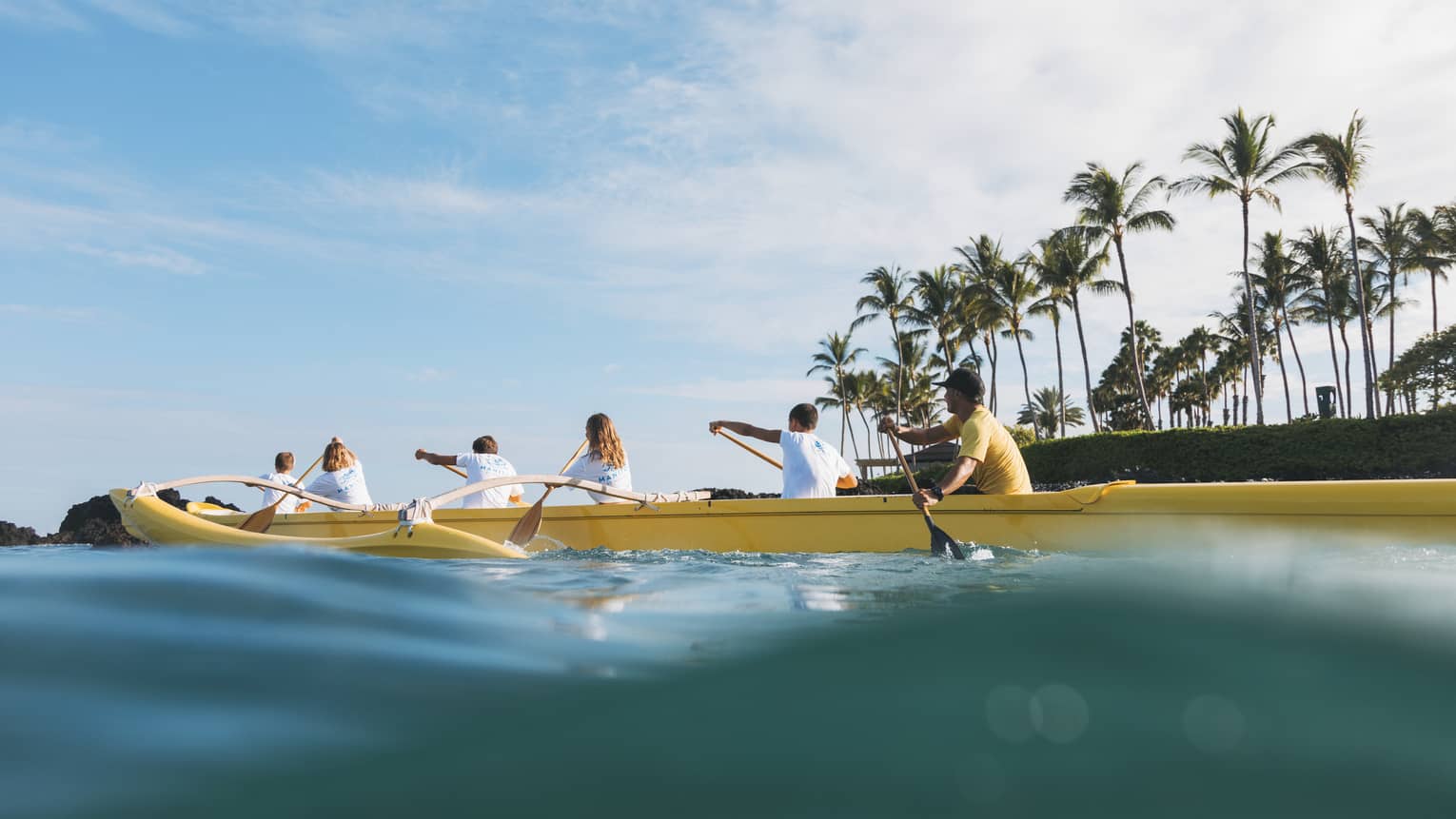 View from water of family and Four Seasons staff paddling outrigger canoe along ocean