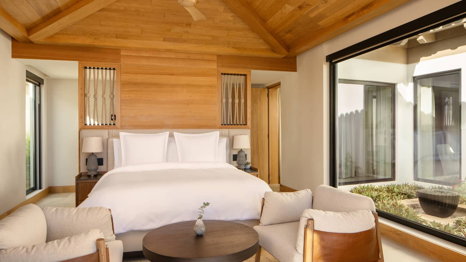 Bedroom with king bed, wooden vaulted ceiling, two arm chairs facing a round coffee table, and a wall of window