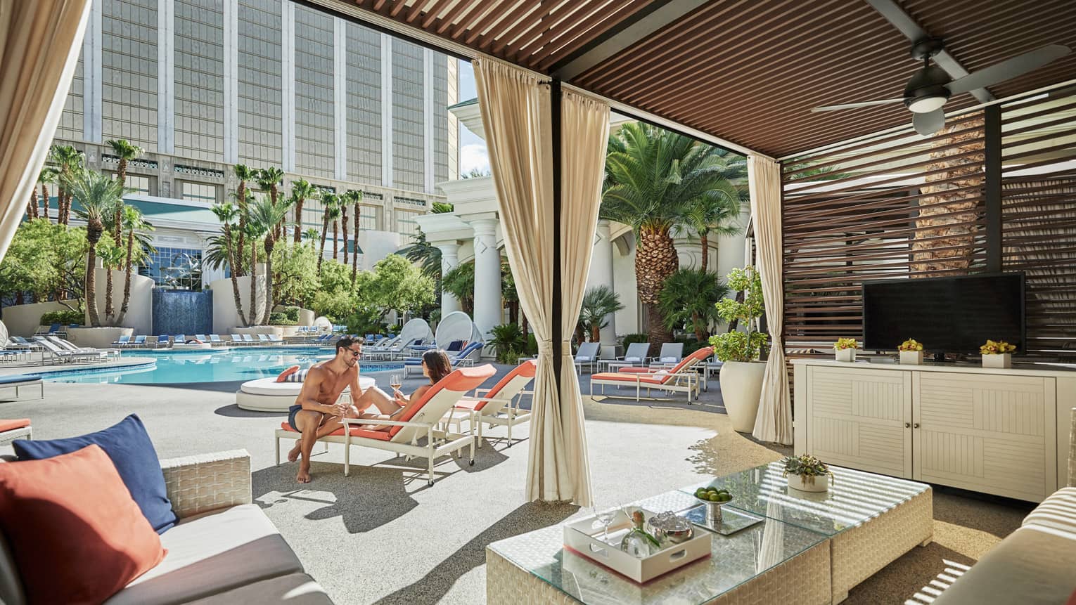 Couple on patio chairs between outdoor swimming pool and cabana seating area 