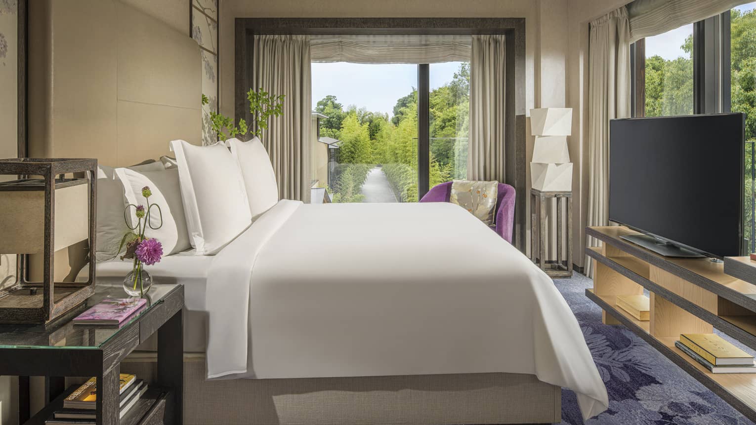 Bright hotel bedroom with a king bed facing TV, side table and a large window overlooking lush greenery