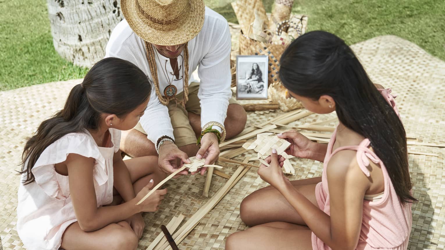 Adult helps two young girls learn how to weave as they sit on a woven blanket