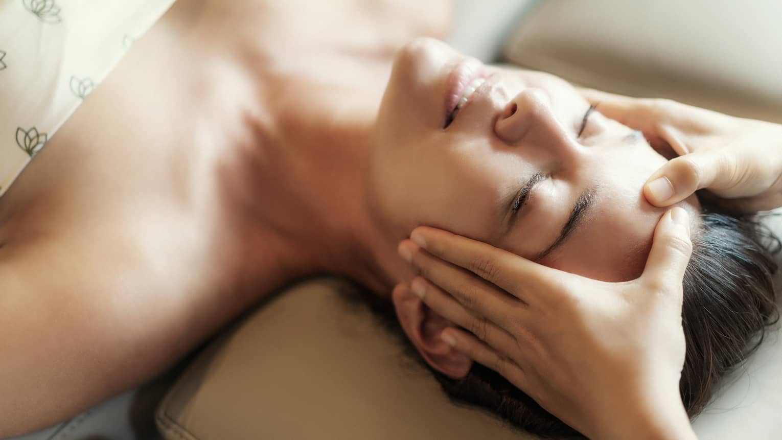 Spa facial, two hands rest on woman's forehead as she closes her eyes, lays under sheet on treatment table