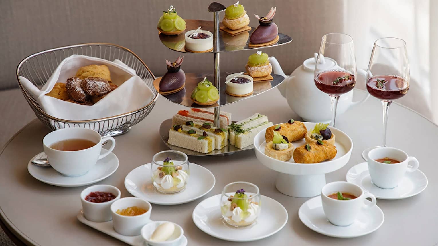 A colourful afternoon tea spread with a tiered tray of sandwiches and petit fours, along with plates of various desserts, served with hot tea and two glasses of red wine