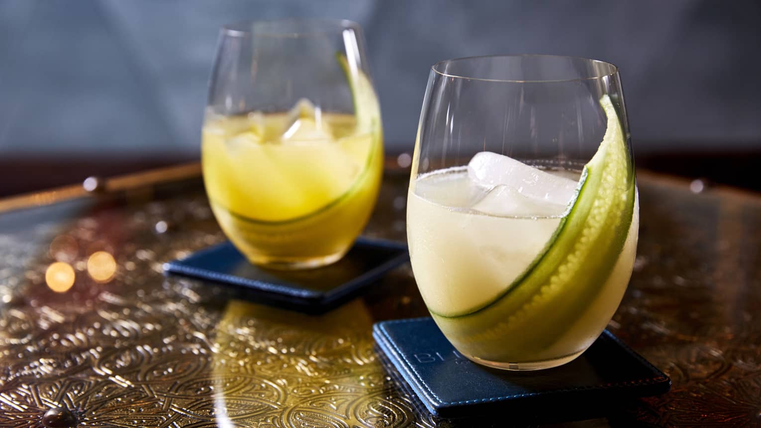 Two yellow beverages in rounded glasses with a green vegetable in the glass.