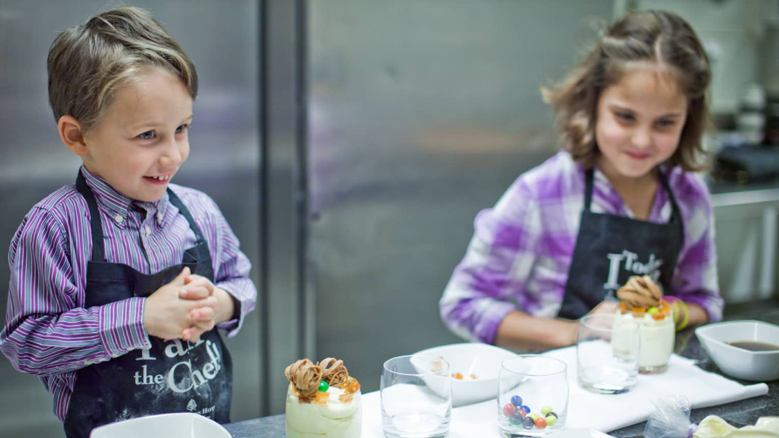 Two young children wearing purple shirts, black aprons prepare desserts at steel counter
