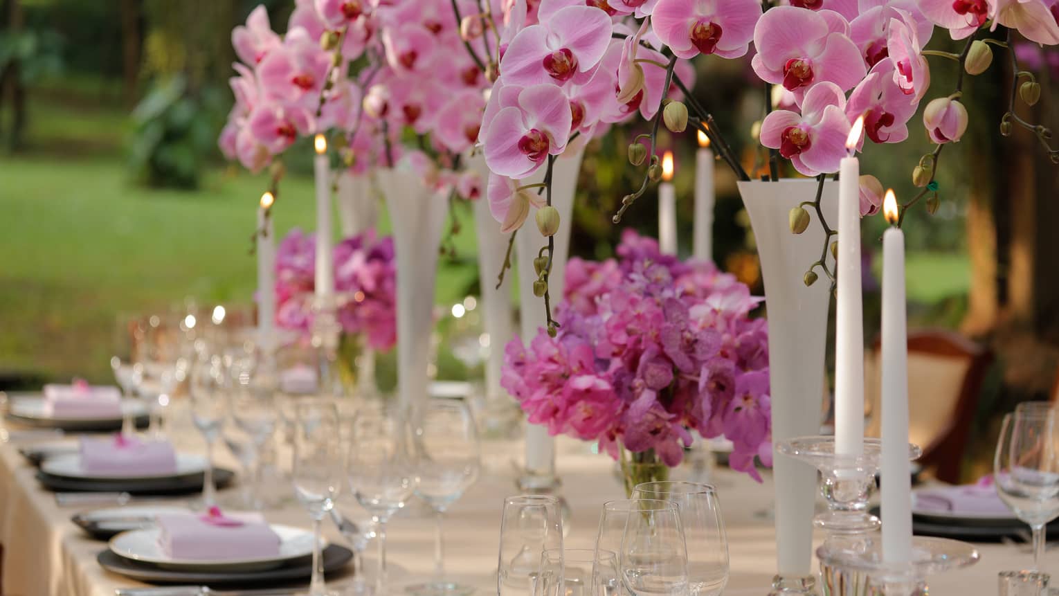 Outdoor dining table with pink flowers in vases, tall white candles 